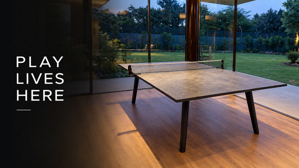 ARGMAC table tennis table with a stone-finish top and black tapered legs, TT net stretched across the middle, glowing warmly indoors beside glass doors opening to the dusky garden.